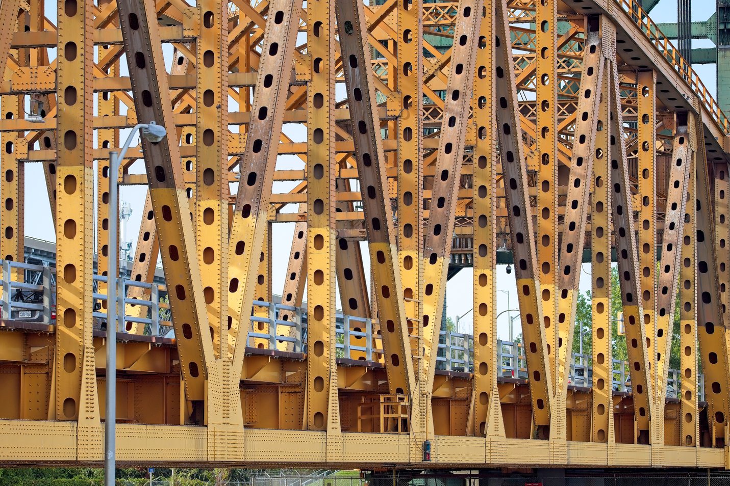 A detailed, close-up view of a massive industrial structure, likely a bridge truss or elevated railway support. The framework is constructed from complex, riveted yellow steel girders arranged in a highly repetitive, lattice pattern, emphasizing the sheer scale and heavy engineering of the material.