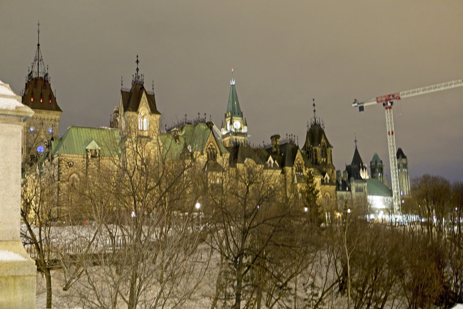 The image captures a large, impressive historical cathedral complex under a dramatic, overcast sky. The foreground is dominated by bare, wintry trees, which frame the massive architecture featuring multiple towers and spires. A construction crane is visible on the right side of the scene.