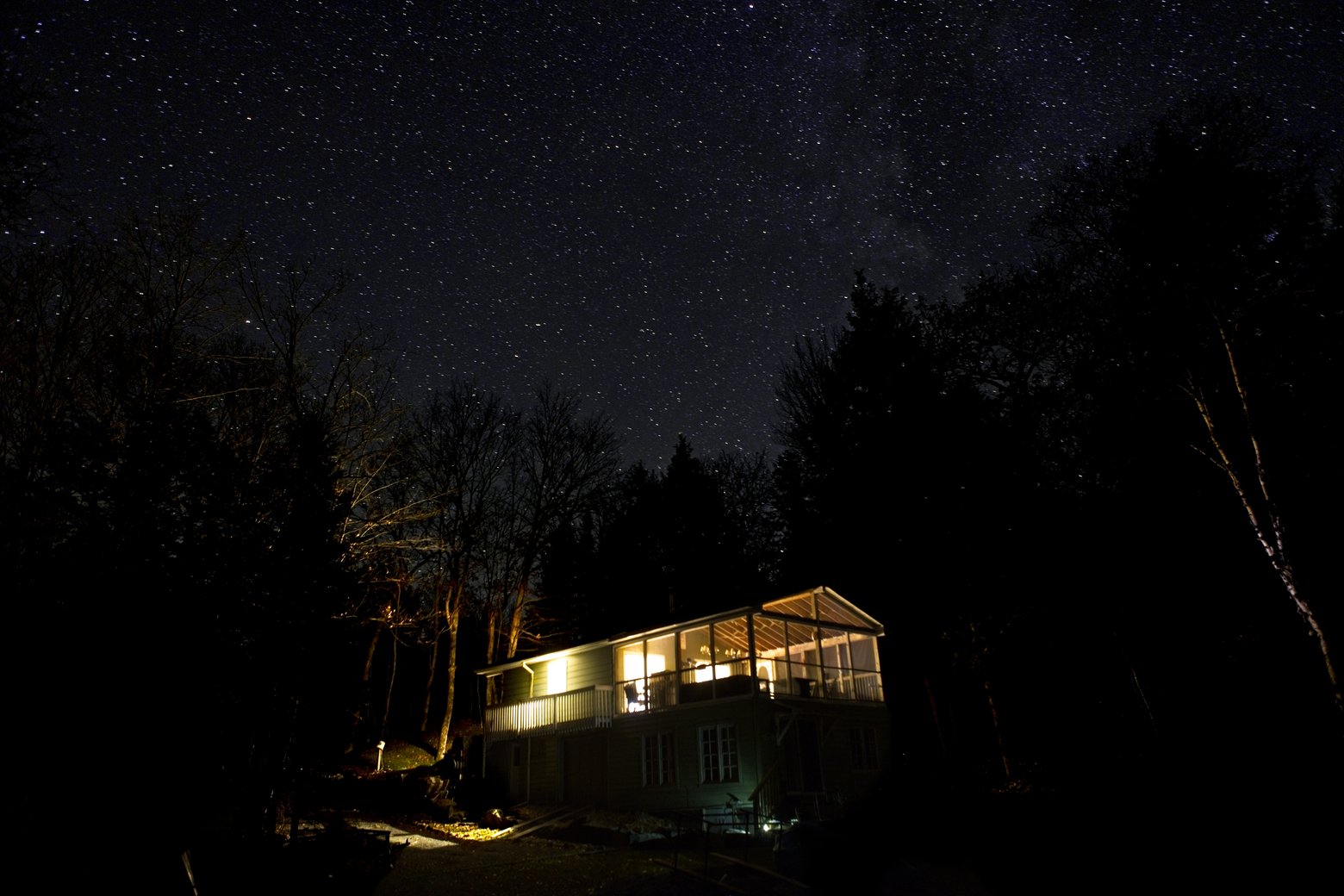 A nocturnal landscape photograph capturing a warmly lit house nestled amongst dense, dark trees, dramatically framed beneath a vast and brilliant star-filled night sky.