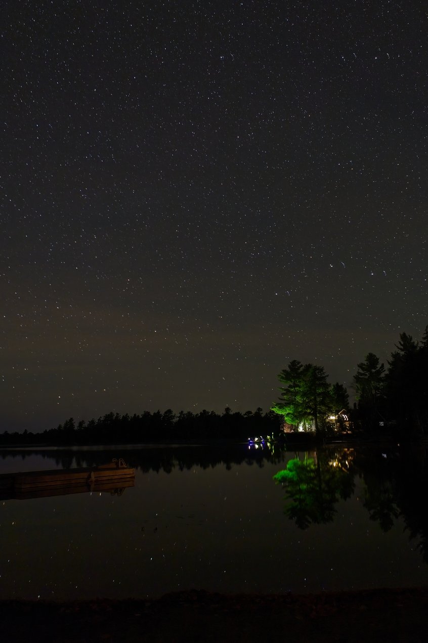 A breathtaking long-exposure photograph capturing a starry night sky reflected in a calm body of water. The scene features a dark, starry expanse above and below, mirroring the celestial details. Along the far shore, silhouetted trees and warm artificial lights provide a contrasting point of civilization and peaceful ambiance to the vast night sky.