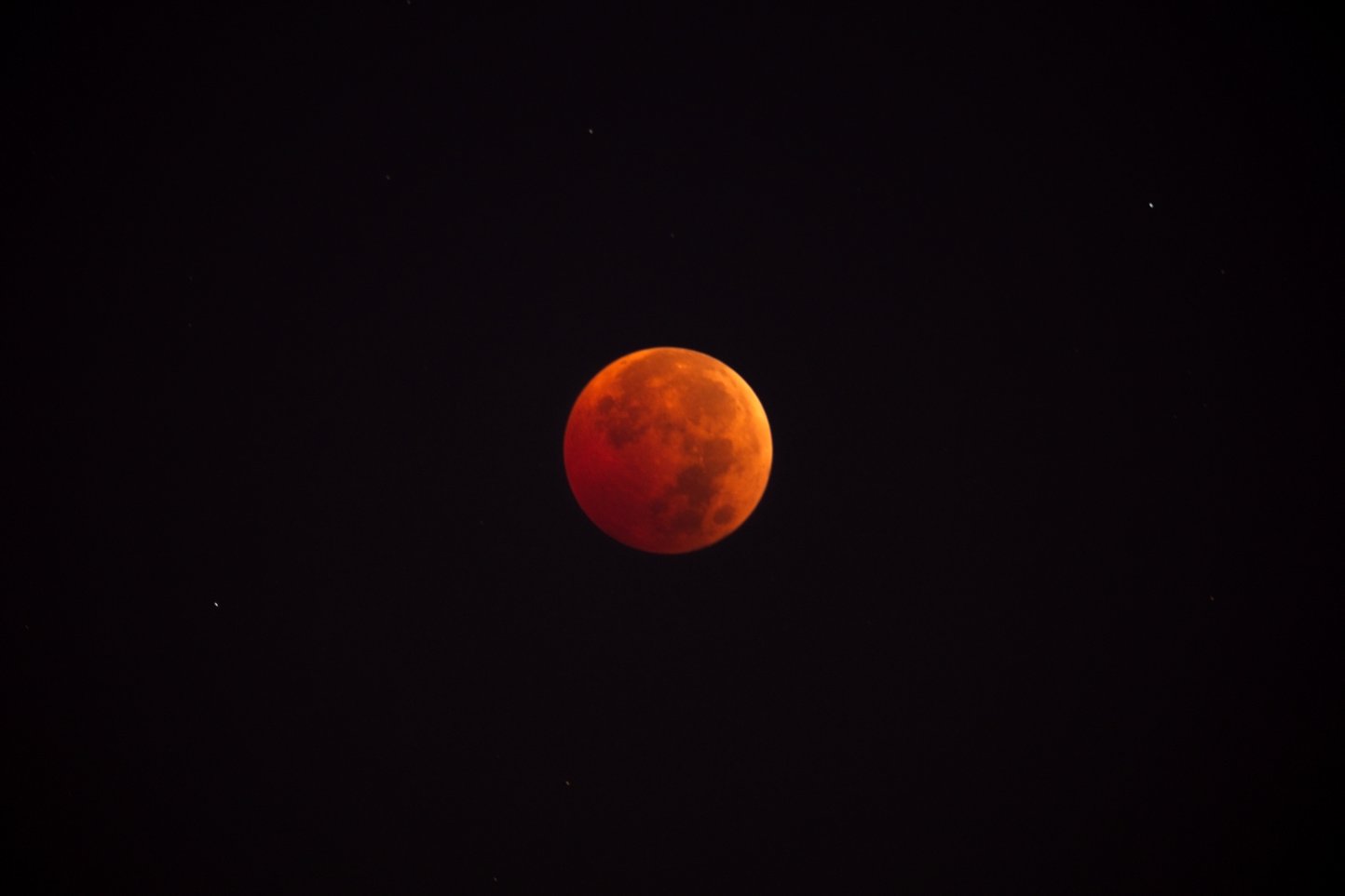 A celestial photograph featuring a reddish-orange Moon, likely captured during a lunar eclipse (Blood Moon), set against a stark, dark black background.
