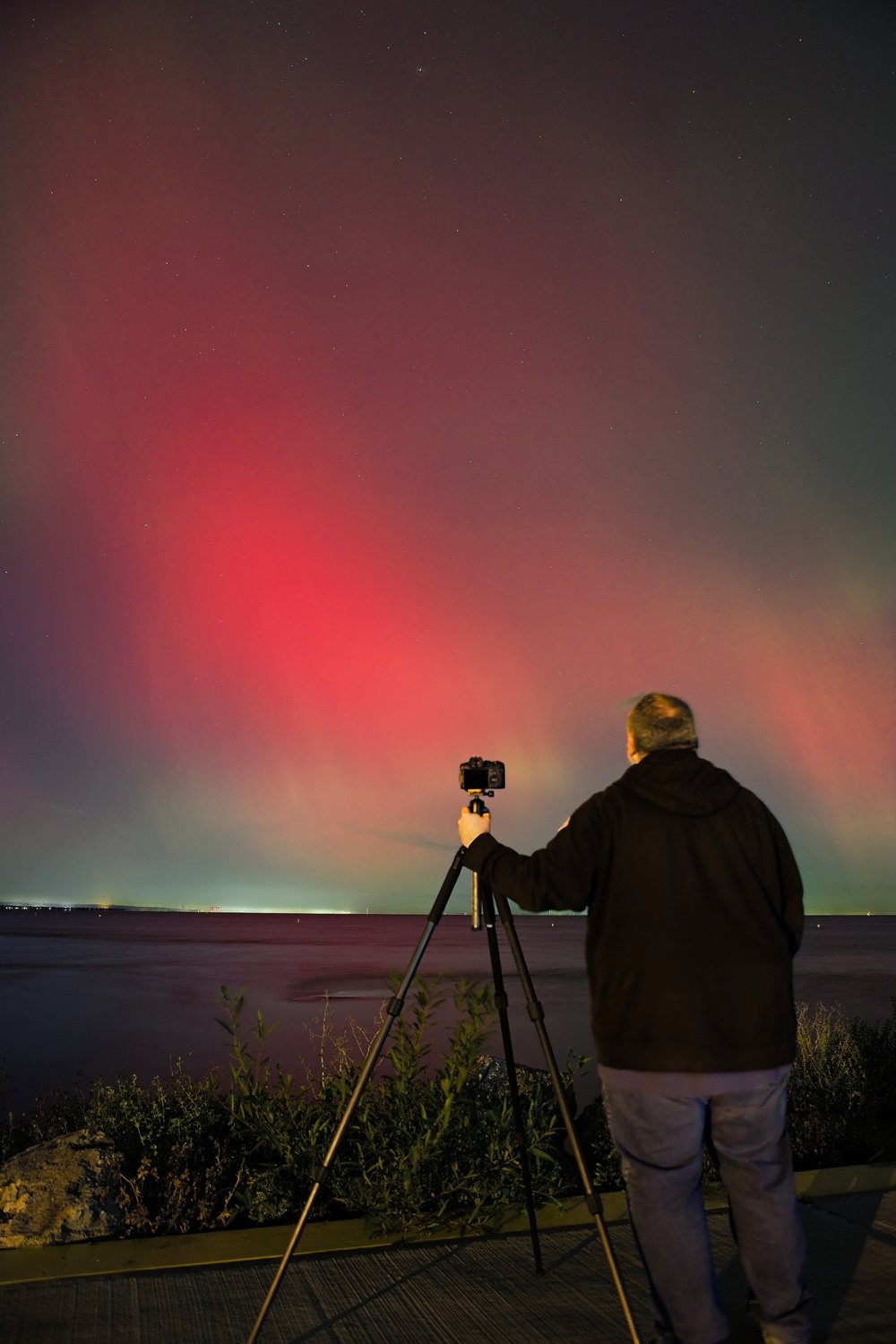A photographer stands by the water at night, setting up equipment to capture the dramatic and vivid red hues of the Aurora Borealis (Northern Lights) in the sky.