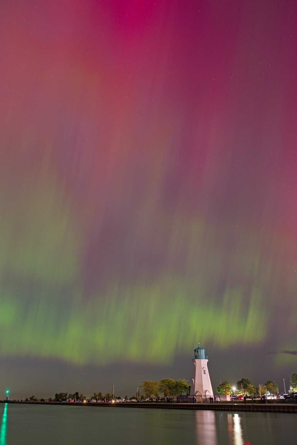 A spectacular nighttime photograph capturing the Aurora Borealis (Northern Lights) displaying vibrant curtains of green and magenta across the sky. In the foreground, a calm body of water reflects the celestial display, while a prominent lighthouse and pier anchor the scene on the horizon, creating a contrast between the ethereal lights and the grounded structure.