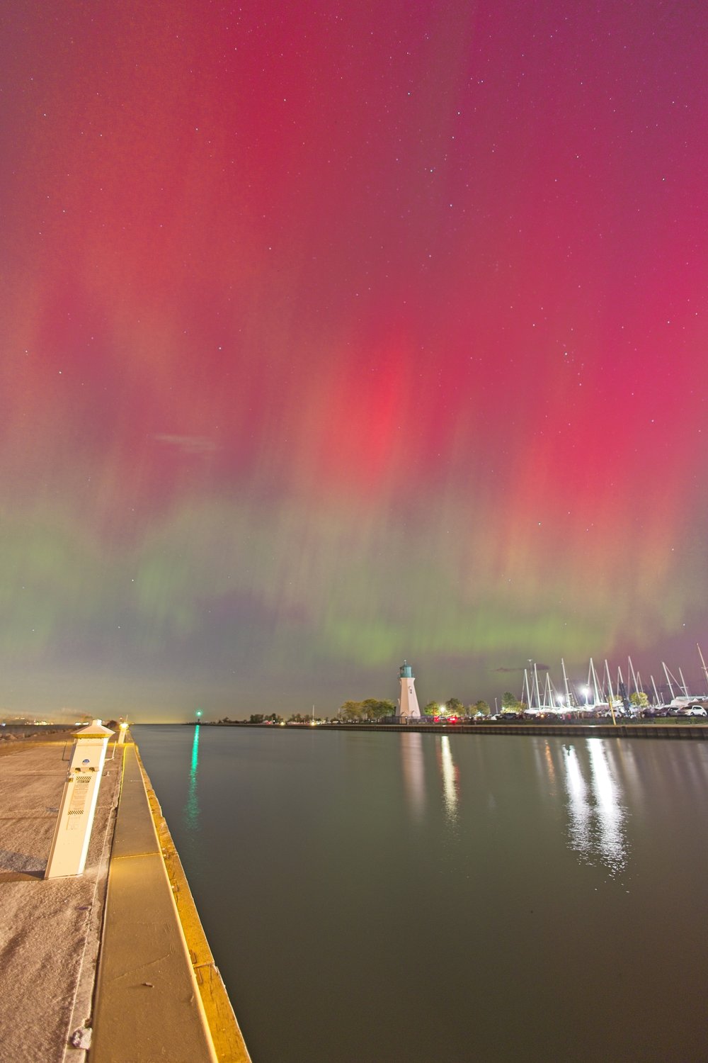 A dramatic long-exposure photograph capturing vibrant red and green auroras (Northern Lights) spanning a night sky. The scene overlooks a calm harbor that perfectly reflects the colorful sky. In the midground, a waterfront promenade leads toward the water, where the far shore features a prominent lighthouse, buildings, and docked sailboats.