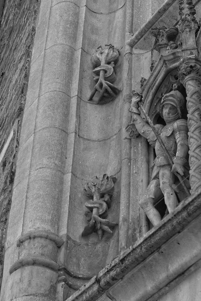 A monochrome photograph showcasing intricate architectural stonework. The image features a prominent pillar structure alongside a decorative niche containing a detailed carved figure. The wall surface is adorned with stylized, organic scrollwork and carvings, typical of historical or gothic architectural detail.