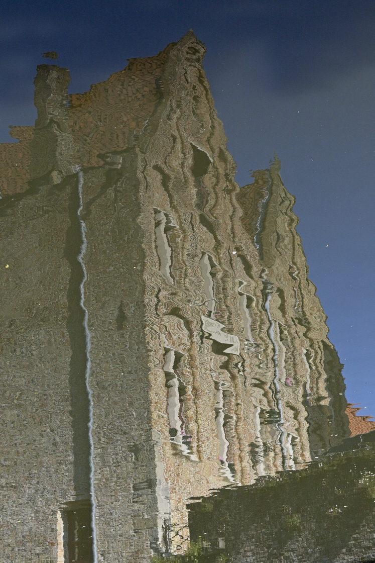 A dramatic view featuring the reflection of massive, aged stone ruins in still water. The deep blue sky and the intricate details of the historical architecture are mirrored, creating a striking contrast between the solid structures and their watery reflection.