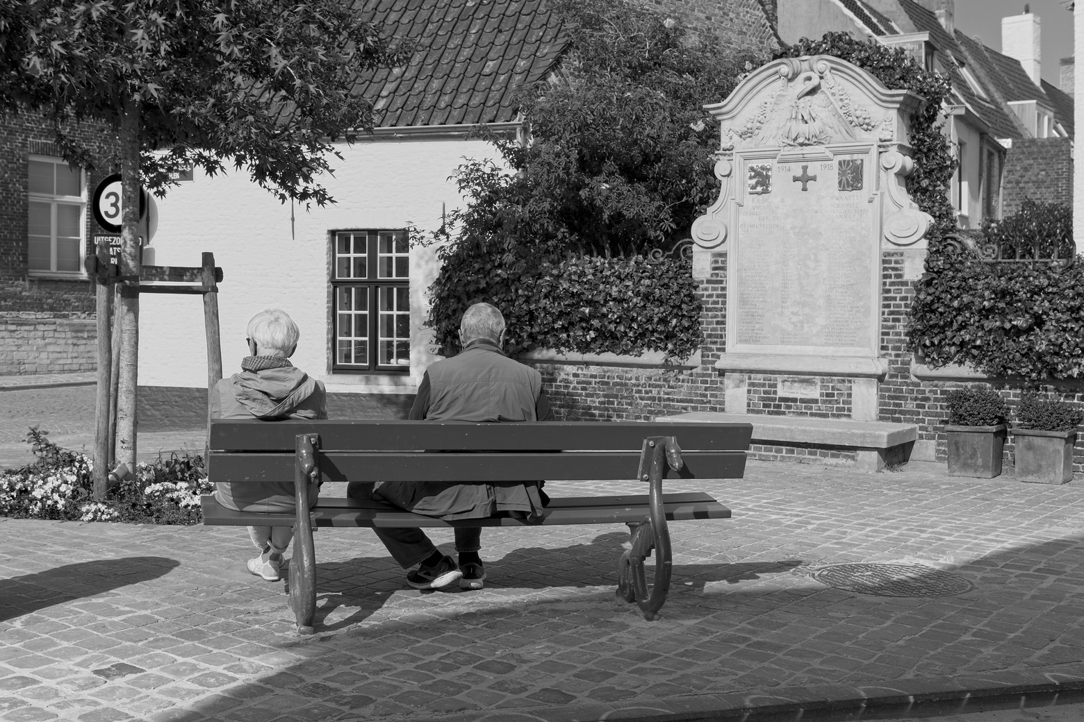 A black and white photograph capturing a tranquil scene in an old town square. Two men are seated on a bench, resting near a large, historical memorial plaque. The background features traditional architecture, lush greenery, and paved cobblestones, giving the image a nostalgic and quiet atmosphere.