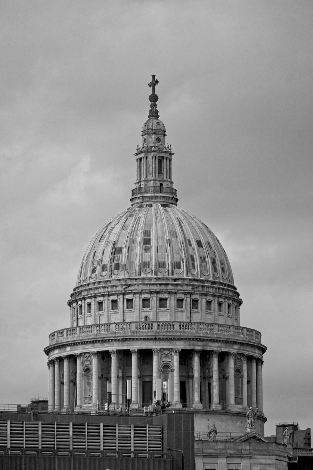 A dramatic black and white architectural photograph featuring St. Paul's Cathedral. The massive domed structure, characterized by classical columns and domes, dominates the frame against a heavily overcast sky. A dark, modern structure is visible in the foreground, creating a contrast between contemporary elements and the historic grandeur of the cathedral.