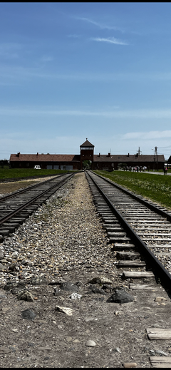 A photograph centered on a pair of railroad tracks receding into the distance, leading toward a classic station building complex. The scene is set on a clear, bright day under a blue sky.