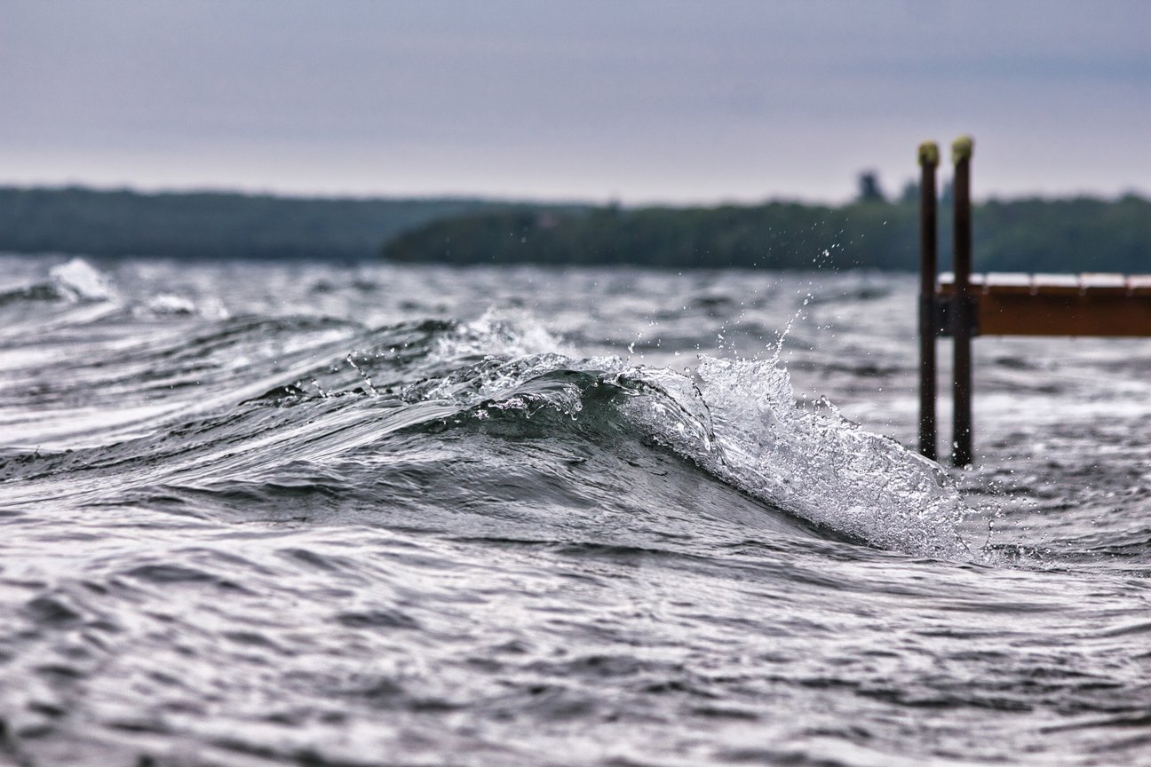 A moody photograph capturing a wave cresting and breaking in choppy, cool-toned water. The foreground focuses on the spray and motion of the wave near visible wooden dock posts, while the background features a hazy, distant shoreline under an overcast sky.