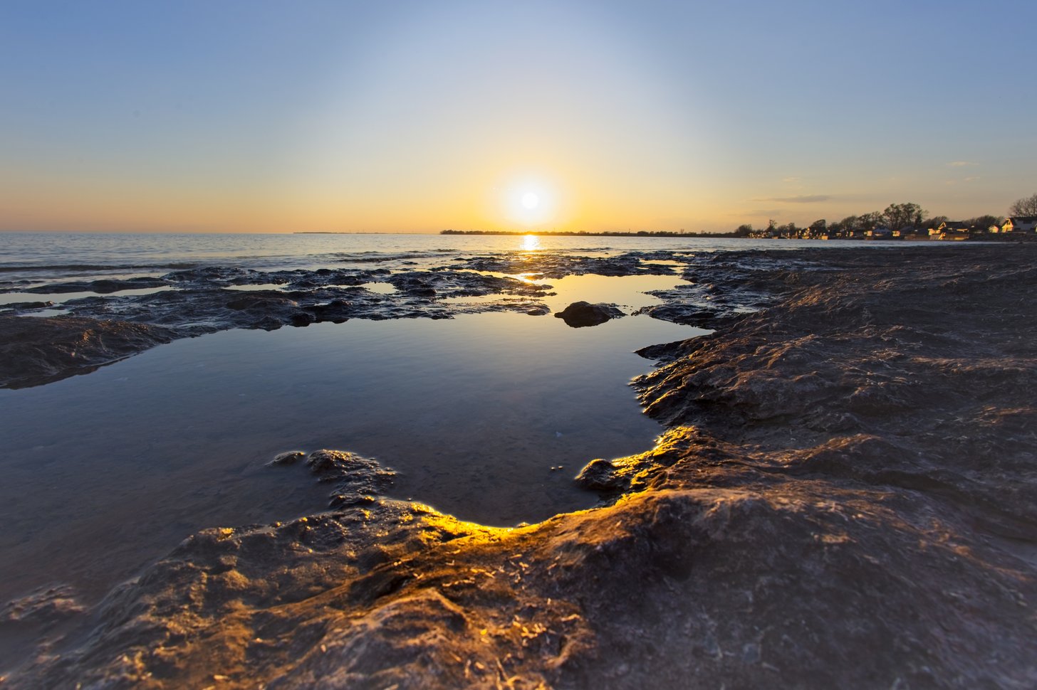 A serene, low-angle photograph capturing a tidal flat area at either sunrise or sunset. The foreground features dark, wet rocks and pools reflecting the warm, golden glow of the sun. The calm horizon and clear sky complete the peaceful coastal scene.