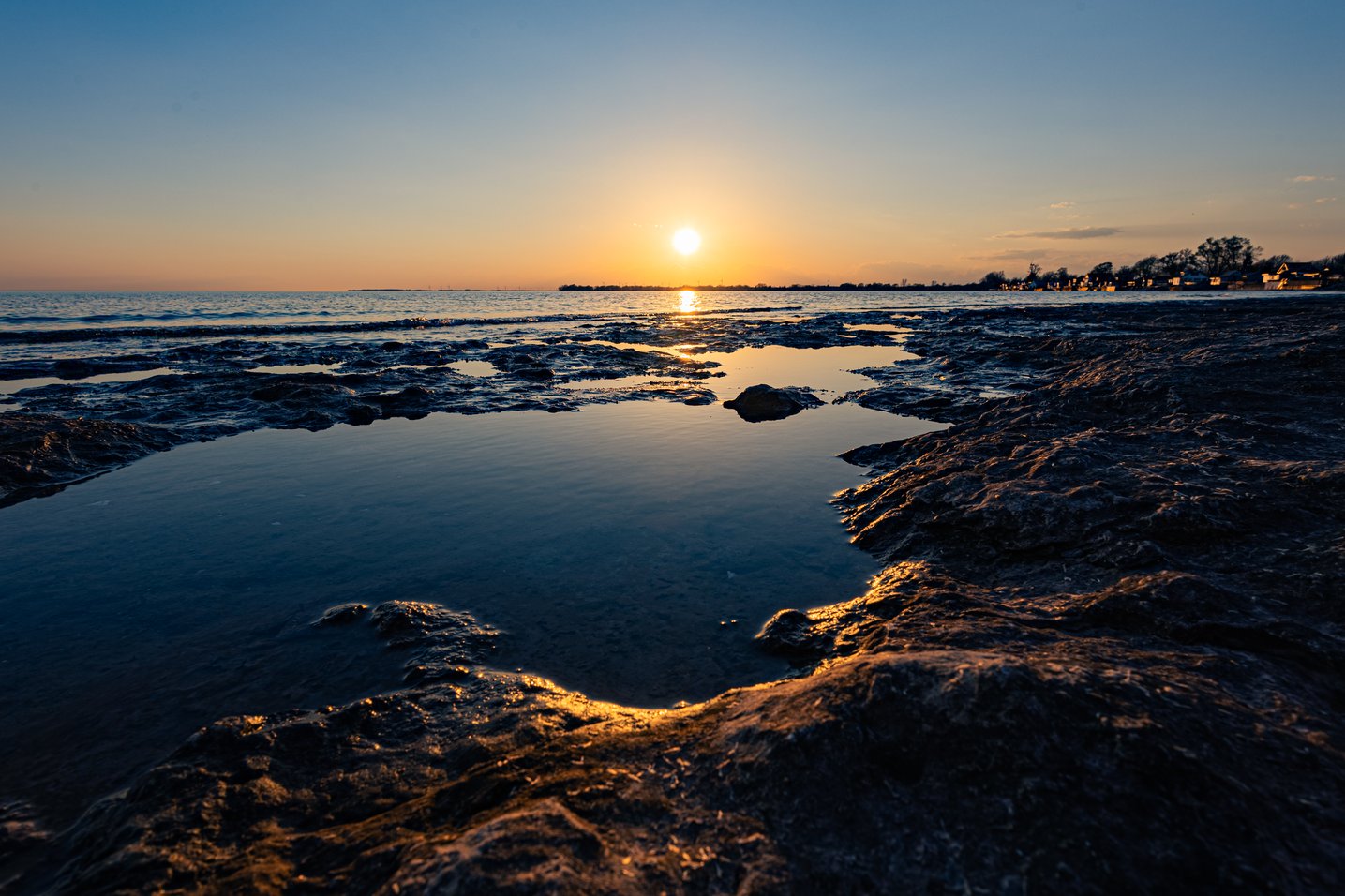 A serene coastal scene during sunrise or sunset, featuring low tide revealing wet mudflats and shallow pools. The golden sun hangs low over the water, casting a brilliant reflection across the water and dark foreground, creating a peaceful and reflective atmosphere.