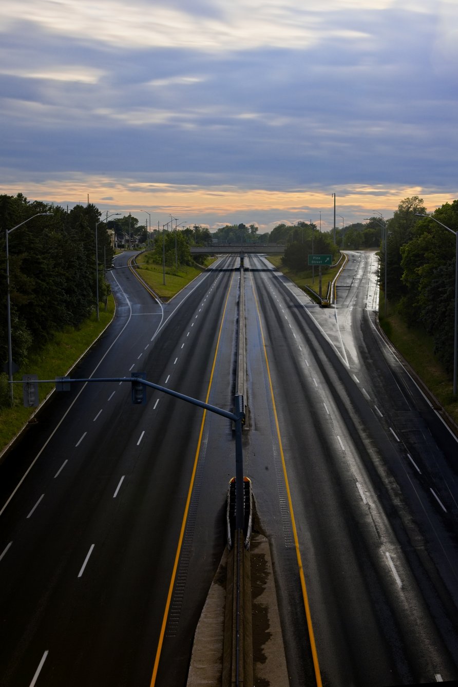 An elevated view of a wide, multi-lane highway wet from recent rain. The road extends into the distance, flanked by lush green trees. The scene is dominated by a cloudy, diffused sky, suggesting early morning or dusk on an overcast day.