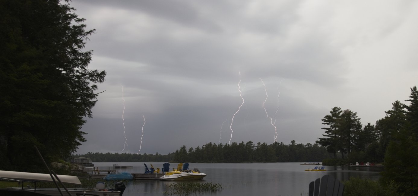 A dramatic, stormy scene over a body of water, dominated by dark, overcast skies. Multiple bright streaks of lightning are visible striking down across the horizon, contrasting with the moody, natural setting.