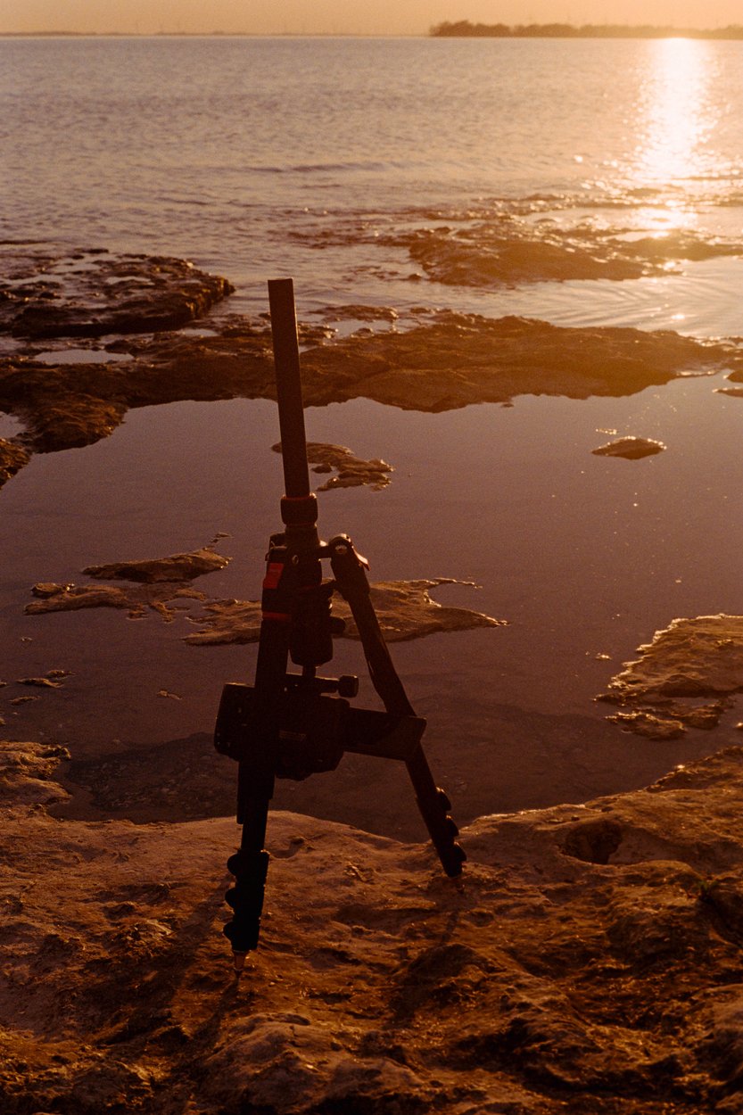 A photograph taken at golden hour on a rocky shoreline. A camera tripod is set up on the damp, dark rocks, facing a calm body of water. The low sun casts a warm, bright reflection across the water's surface, creating a tranquil and dramatic coastal scene.